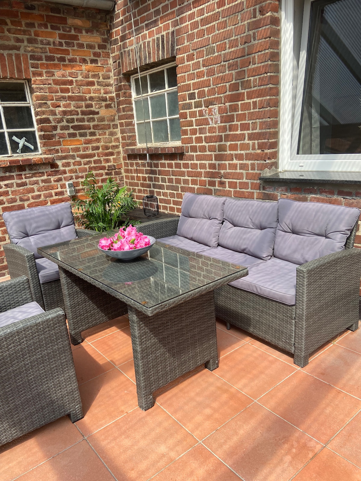 A comfortable outdoor seating area features a glass-top table surrounded by gray wicker furniture, including a sofa and two chairs. Bright pink flowers are displayed in a bowl at the center of the table. Exposed brick walls and tiled flooring provide a rustic backdrop.
