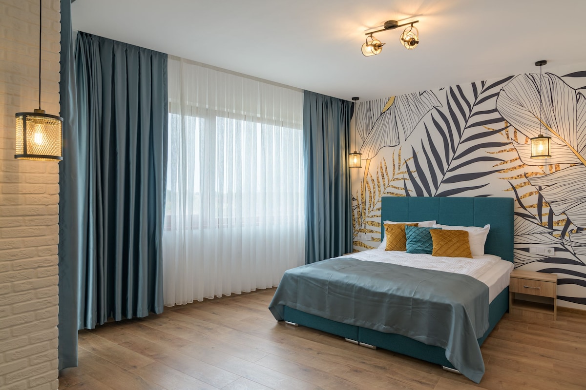 A spacious sleeping area features a calming blue and white color palette. A large bed with decorative pillows is positioned against a patterned wall. Soft natural light filters through sheer curtains, and wall sconces provide additional illumination.