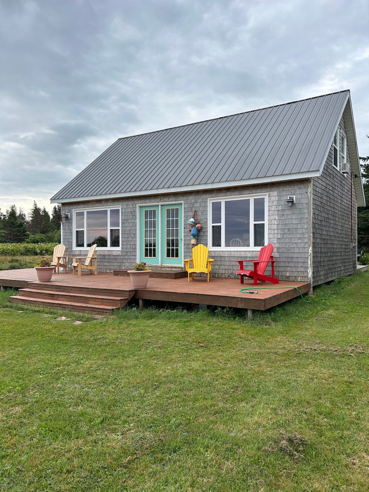 A charming cottage with a grey shingle exterior features a green front door and multiple large windows. A spacious wooden deck is adorned with colorful adirondack chairs in yellow and red, surrounded by grassy areas and a backdrop of trees.
