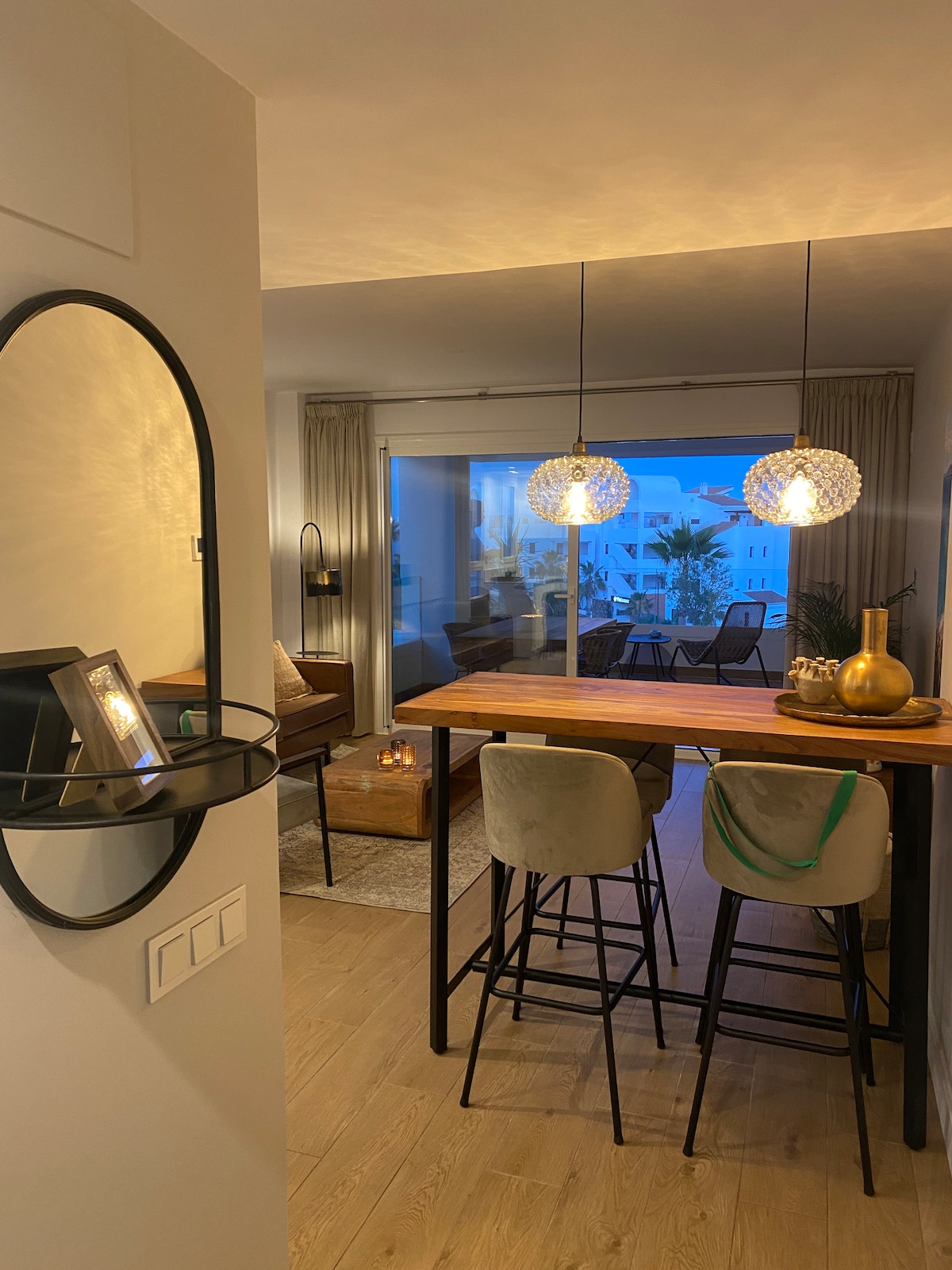 A stylish dining area features a wooden table illuminated by two pendant lights. Barstools provide seating, and a mirror hangs on the wall near the entrance. Large glass doors open to a balcony, adding natural light to the welcoming space.