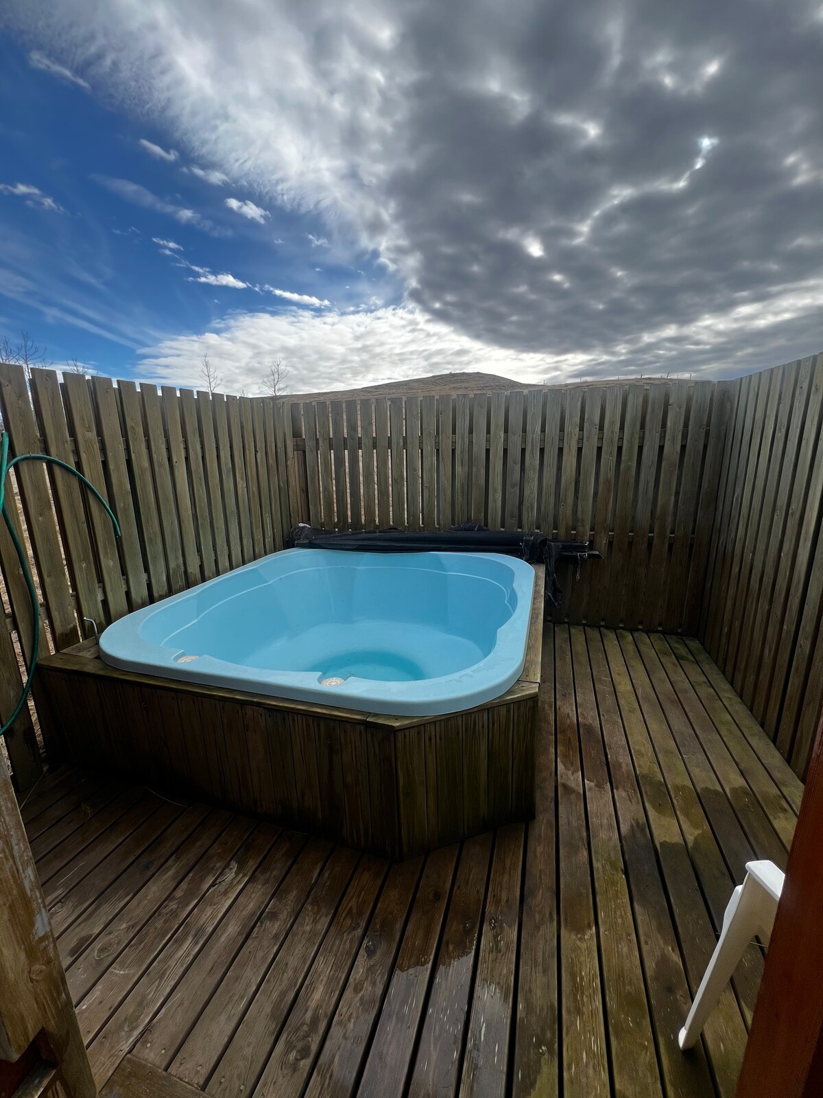 A hot tub is surrounded by wooden decking within a private fenced area. The structure features tall wooden slats that create a sense of seclusion. Expansive clouds are visible in the sky above, adding to the outdoor ambiance.