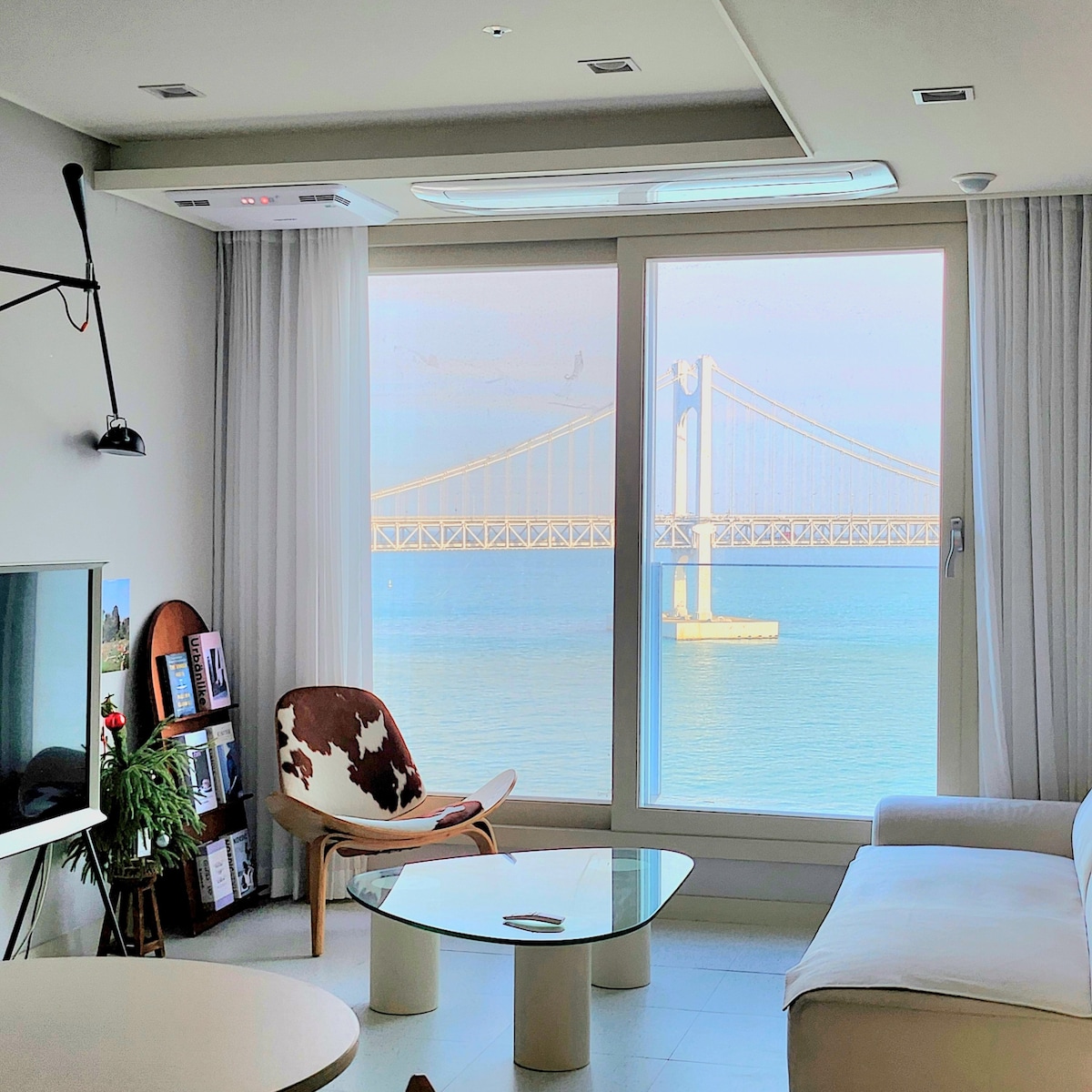 A well-lit living space features a large window showcasing a scenic view of a bridge over water. A white sofa and a stylish chair are positioned near a glass coffee table, contributing to the open and airy atmosphere. Natural light fills the area.