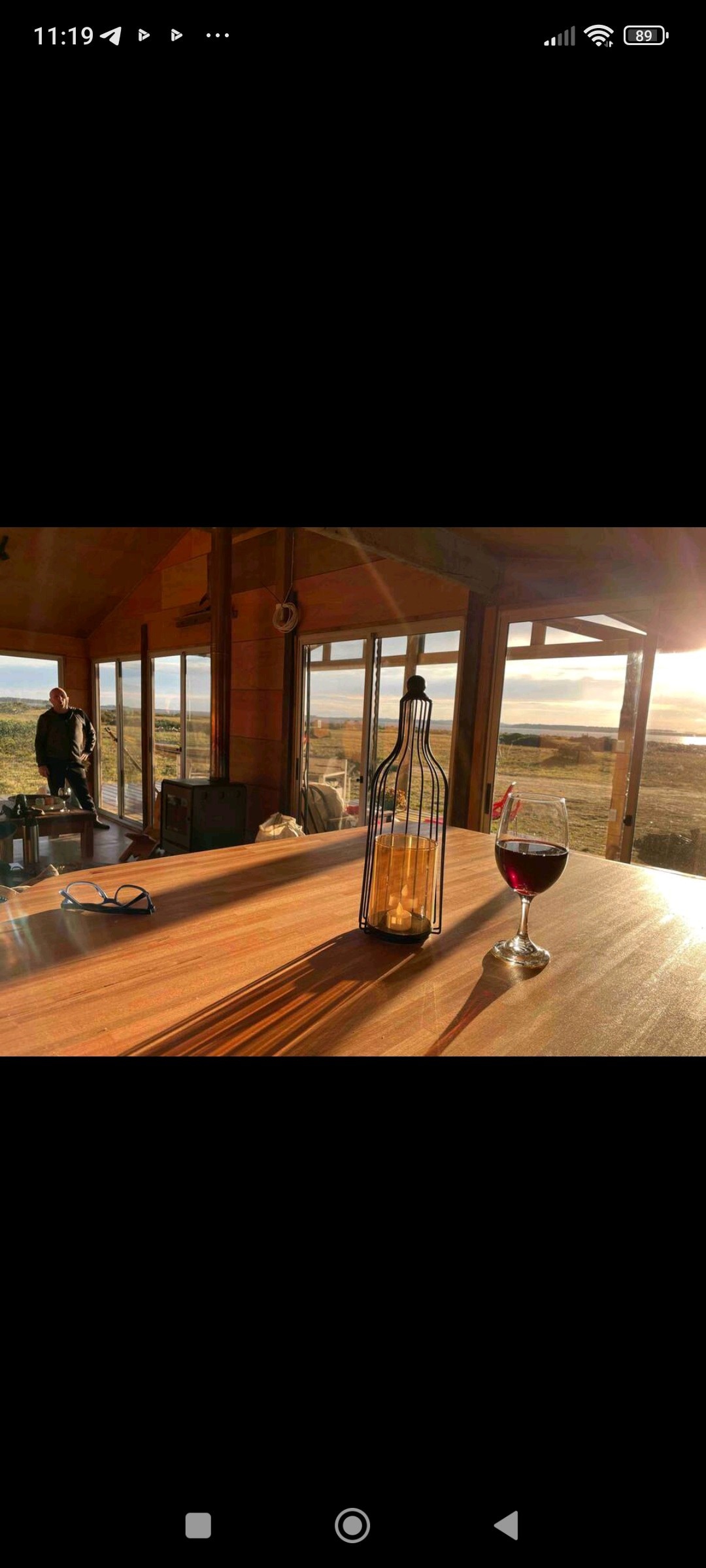 A wooden table is set with a wine glass filled with red wine and an empty bottle, casting long shadows in the warm light. Large windows reveal a scenic view of the landscape, enhancing the serene ambiance of the space.