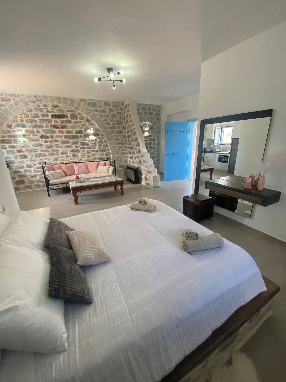 A spacious sleeping area features a large bed adorned with neutral linens. A stone wall serves as a striking backdrop, while a cozy seating area with soft cushions invites relaxation. A wooden table and a large mirror are positioned nearby, enhancing the room's overall charm.