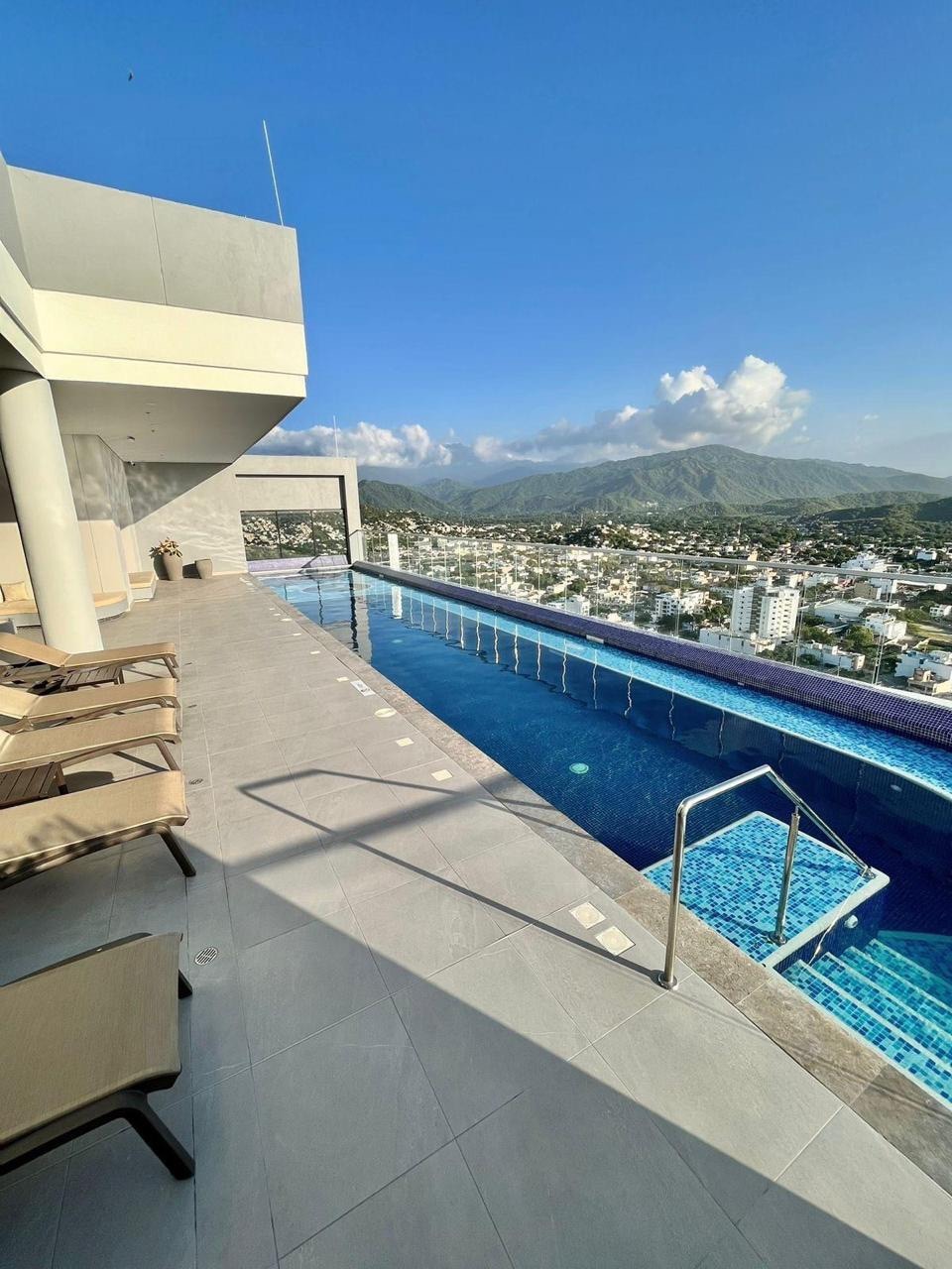 An infinity pool extends along a rooftop terrace, offering expansive views of the surrounding mountains and cityscape. Sun loungers are placed nearby, providing a comfortable space to relax under the clear blue sky. The pool's edge blends seamlessly with the horizon.