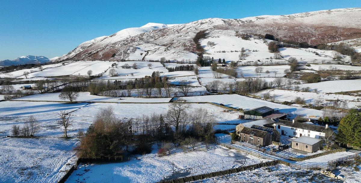 A scenic view of a snow-covered landscape showcases rolling hills and trees dotted throughout. The property is positioned along a quiet rural area, with a collection of buildings visible, emphasizing the peaceful charm of the natural surroundings.