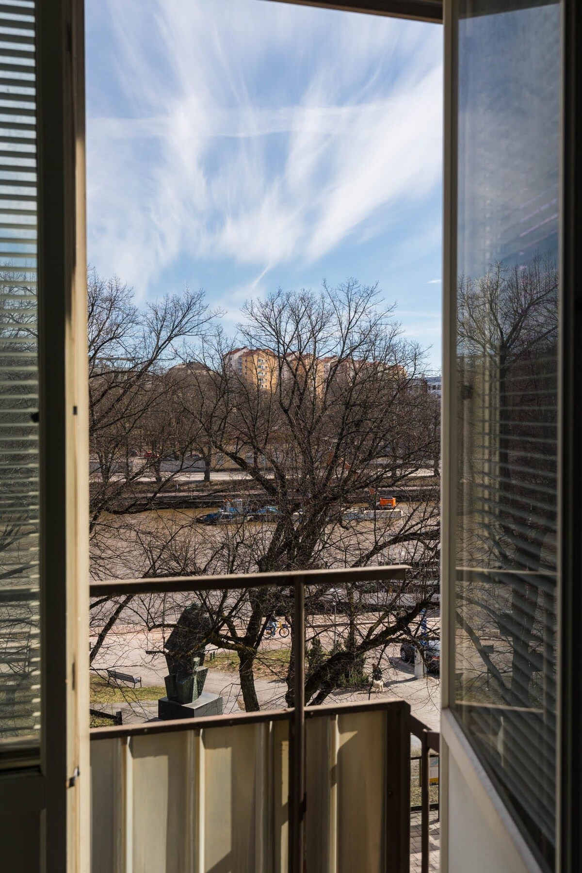 A view from an open balcony showcases a clear sky with soft, wispy clouds above. The scene includes bare trees in the foreground, indicating a tranquil outdoor area, and distant buildings visible in the background.
