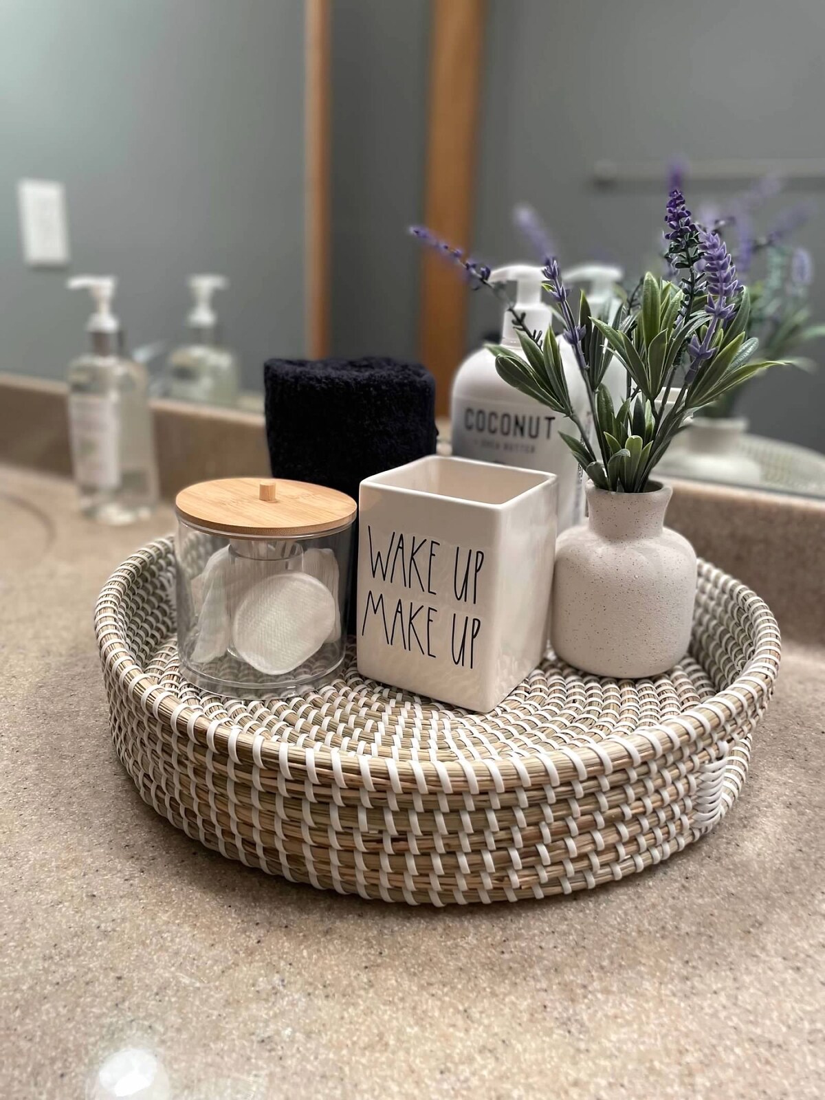 A round woven tray is positioned on a countertop, featuring a container labeled 'WAKE UP MAKE UP' and a glass jar. A small potted plant adds a touch of greenery, alongside a black towel, with additional toiletries visible in the background.