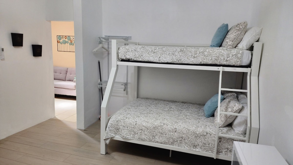 A private room is furnished with a full-twin bunk bed featuring patterned bedding and soft pillows. Light-colored walls provide a bright atmosphere, while a clothing rack offers additional storage. A shared lounge area is visible in the background, enhancing accessibility.