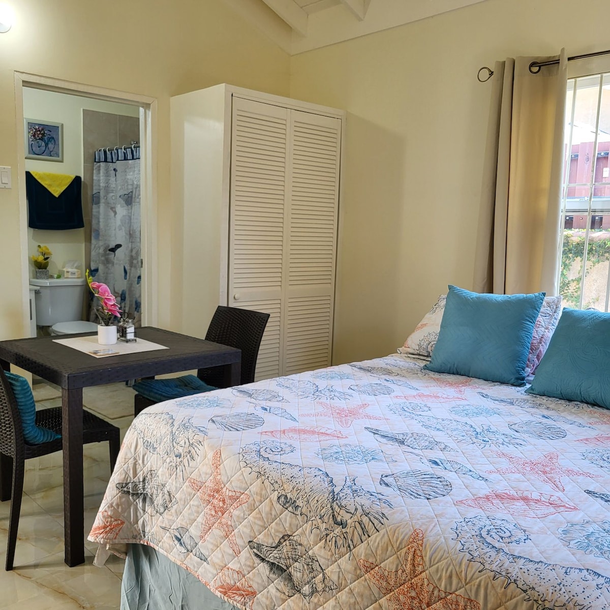 A spacious sleeping area features a comfortable bed adorned with a light quilt and multiple blue cushions. A small dining table with two chairs is positioned nearby, while a closet can be seen in the background. Soft light enters through a window, illuminating the room.