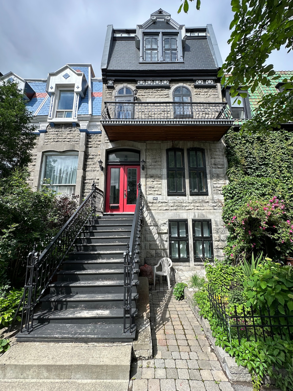 A stone facade stands prominently, featuring a steep staircase leading to an entrance with a bright red door. Lush greenery surrounds the building, with flower beds visible along the path. Architectural details reflect a historic charm, enhanced by decorative elements on the upper levels.
