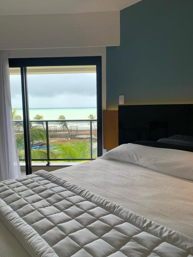 A modern bedroom showcases a spacious bed with an elegant quilted blanket. Large glass doors provide access to a balcony, offering a view of the ocean and surrounding palm trees. Natural light fills the room, highlighting the calming color scheme.