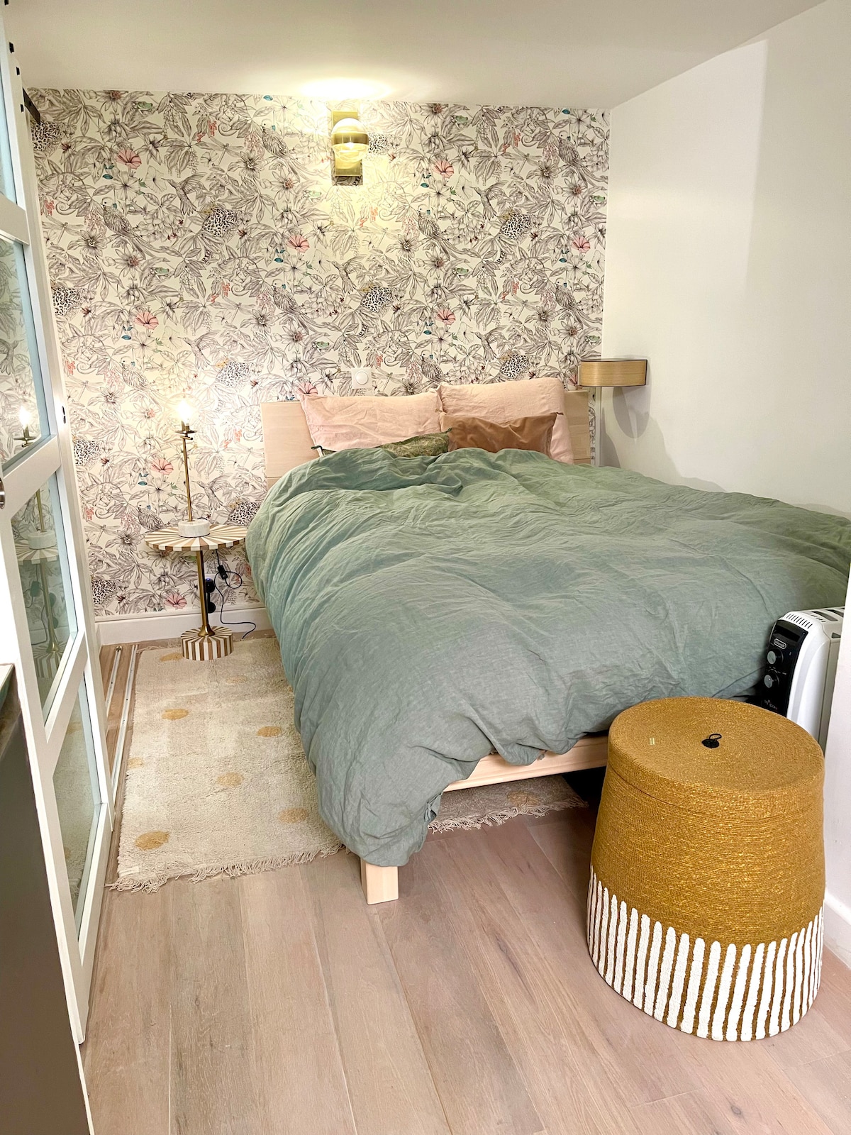 A compact sleeping area features a spacious bed draped with a soft, green duvet. Coordinated pillows enhance the bedding. A bedside table holds a lamp, and a textural ottoman adds comfort. A patterned wallpaper creates a serene backdrop, while natural light filters into the room.