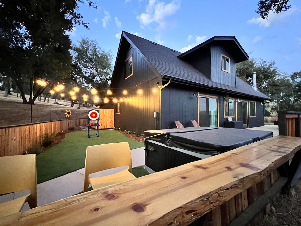 A modern cabin exterior is showcased, featuring a cozy patio area with string lights overhead. A hot tub is positioned beside yellow chairs, while a well-maintained lawn area provides green accents. The structure is framed by trees, enhancing the tranquil outdoor setting.