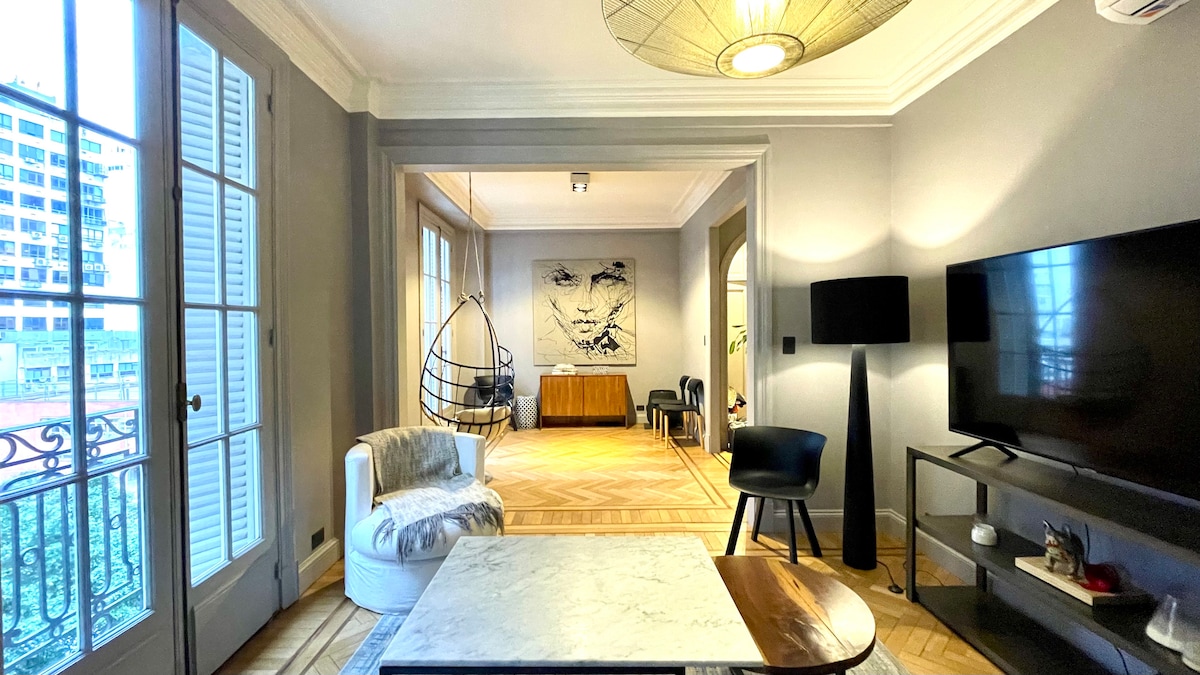 A spacious living area is highlighted by elegant, neutral tones. A large window with shuttered doors allows natural light to fill the room. A modern television is positioned on a sleek cabinet, while a stylish chair and unique hanging seat provide relaxed seating options.