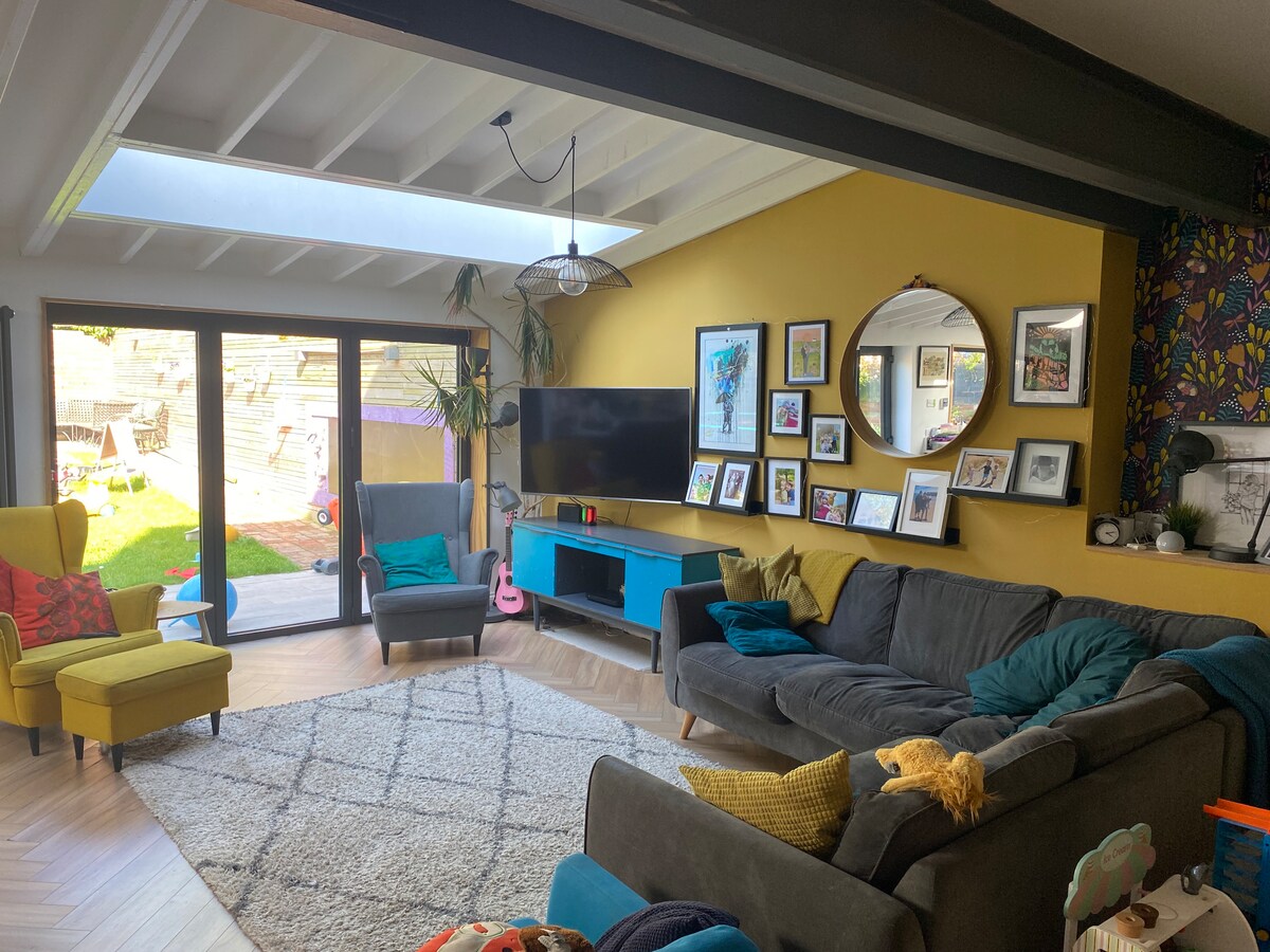 A bright living area is displayed, featuring a spacious grey sofa adorned with colorful pillows. A round mirror hangs above a vibrant blue cabinet, while framed artwork lines the walls. Natural light floods in through skylights and glass doors, enhancing the contemporary decor.