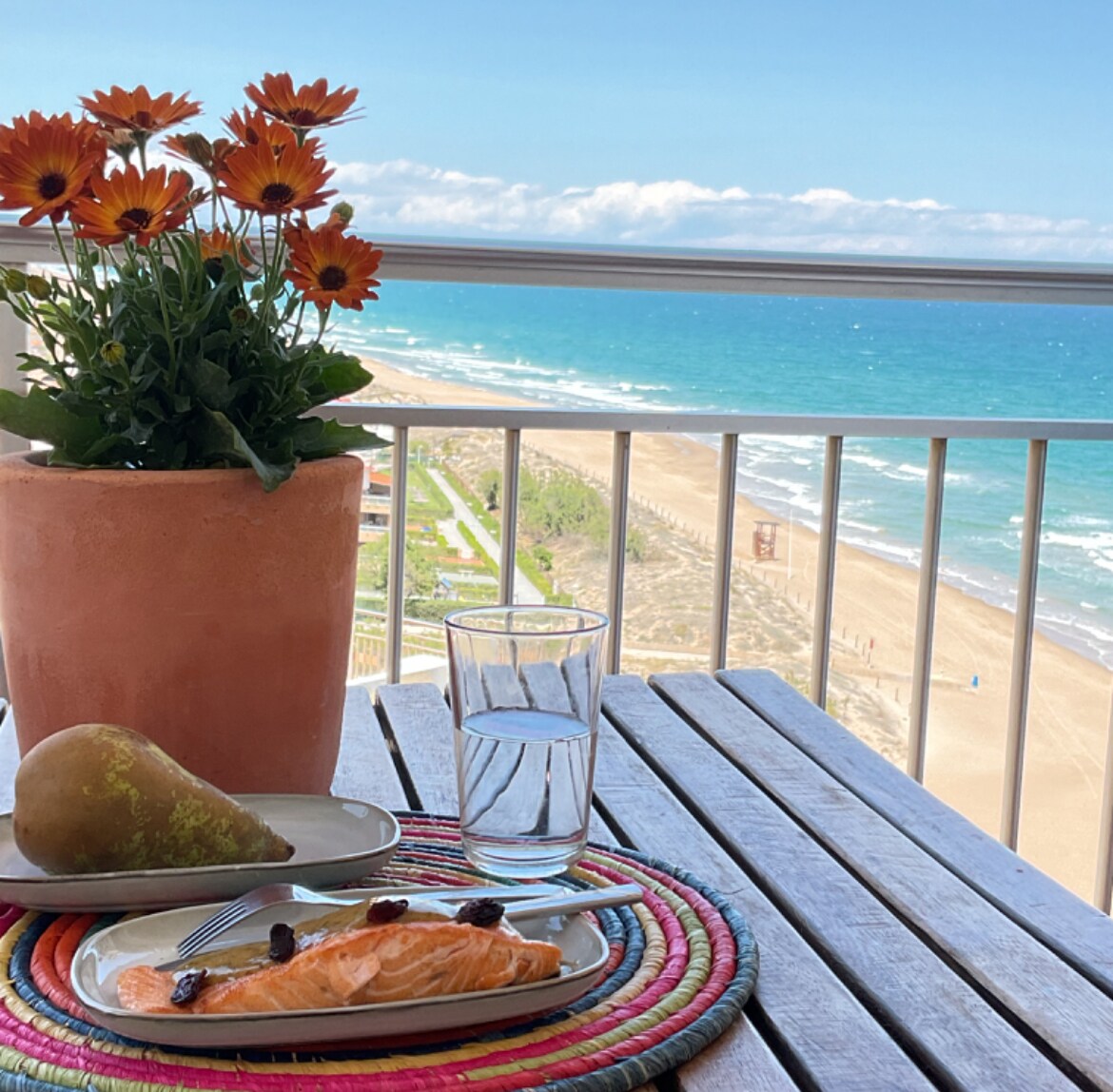 A dining table is set on a balcony, featuring a vibrant arrangement of orange flowers in a terracotta pot. A plate of salmon is placed on a colorful woven mat alongside a glass of water, with expansive views of the sandy beach and turquoise sea in the background.