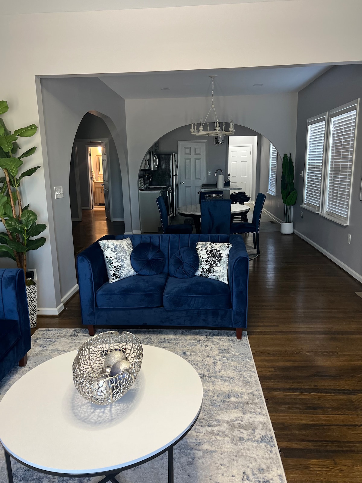 A cozy living area presents a plush, navy blue sofa with decorative cushions, complemented by a round coffee table at the center. In the background, an open dining space is visible, featuring a chandelier and dark wood dining chairs around a table. Natural light enters through several windows.