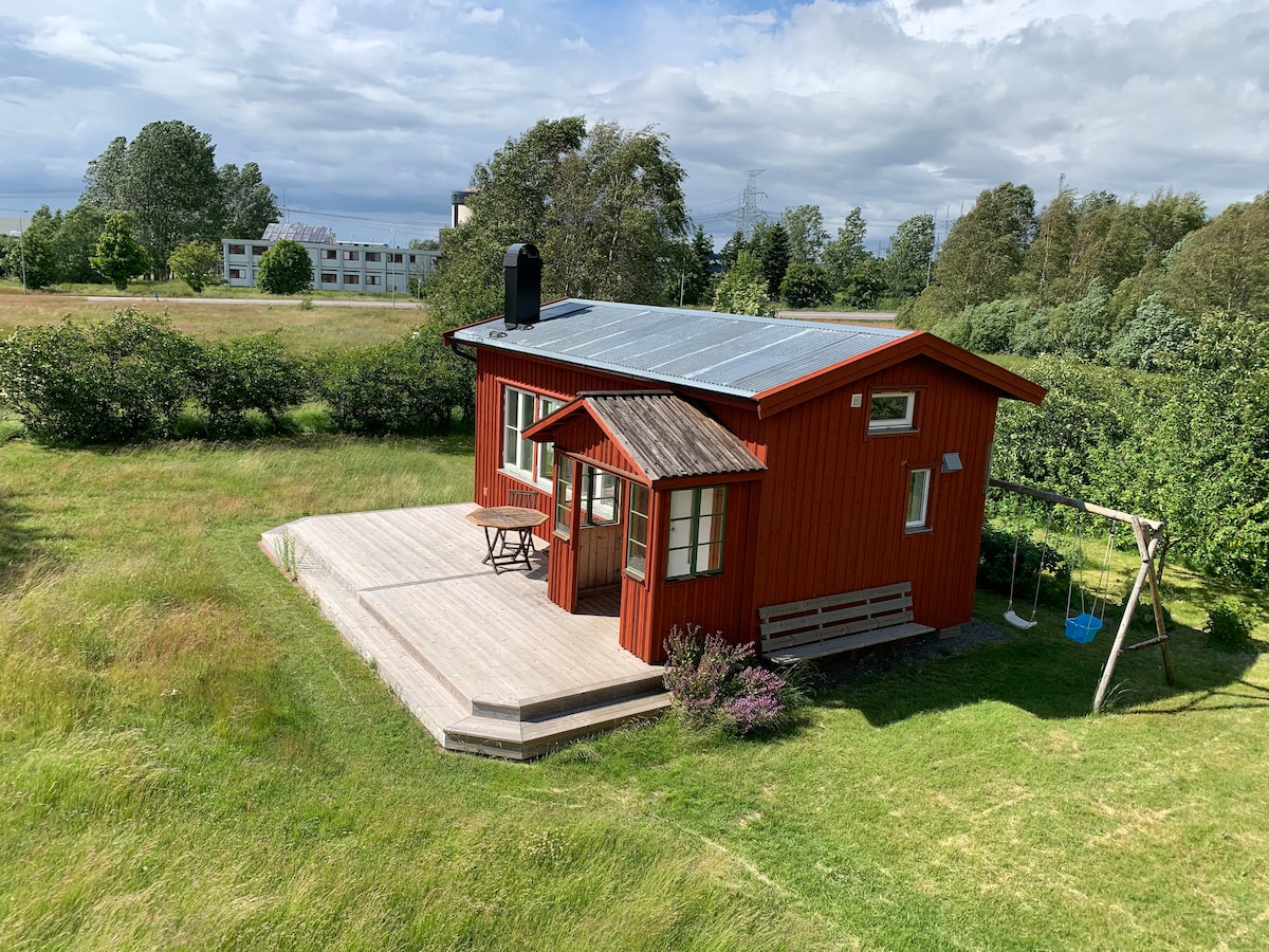 A red wooden cottage is set within a grassy landscape, featuring a deck with outdoor seating. Solar panels are installed on the roof. Surrounding greenery adds to the serene environment, with a swing set visible to one side.
