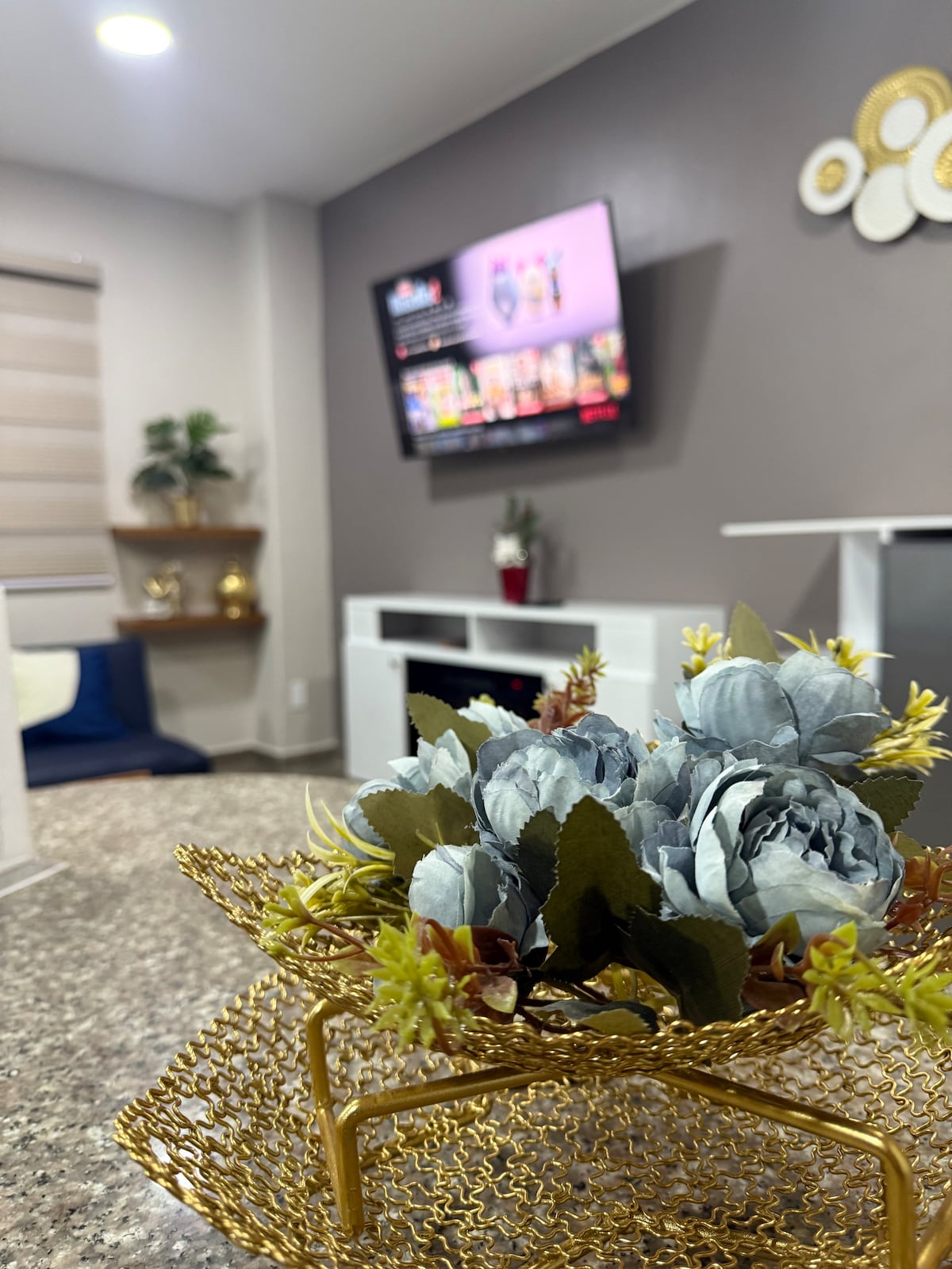 A living area features a modern television mounted on a wall above a white cabinet. A decorative golden basket holds artificial flowers at the forefront. Soft furnishings are visible in the background, along with plants and a light-filtering window treatment.