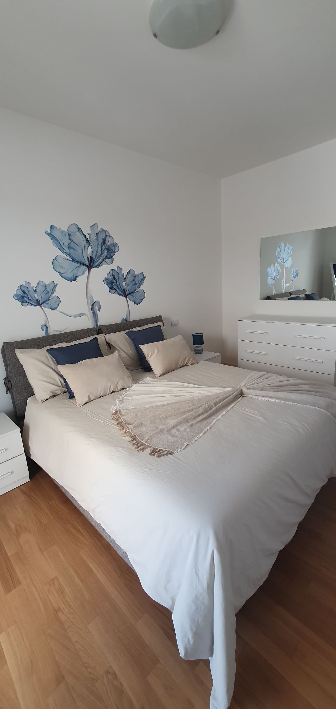 The bedroom features a spacious bed with soft linens and decorative pillows. A floral mural adds color to the wall. A mirror and dresser are positioned nearby, with bedside tables on each side, creating a functional and inviting layout.