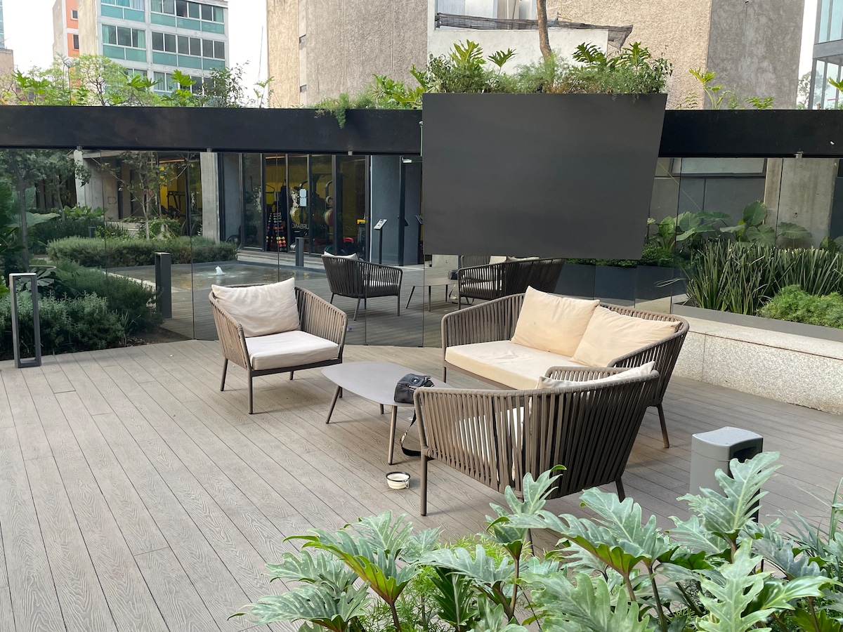 An outdoor lounge area features a pair of minimalist armchairs and a spacious sofa, arranged around a sleek coffee table. Lush greenery surrounds the wooden deck, and large windows reflect the modern architecture of the nearby building.
