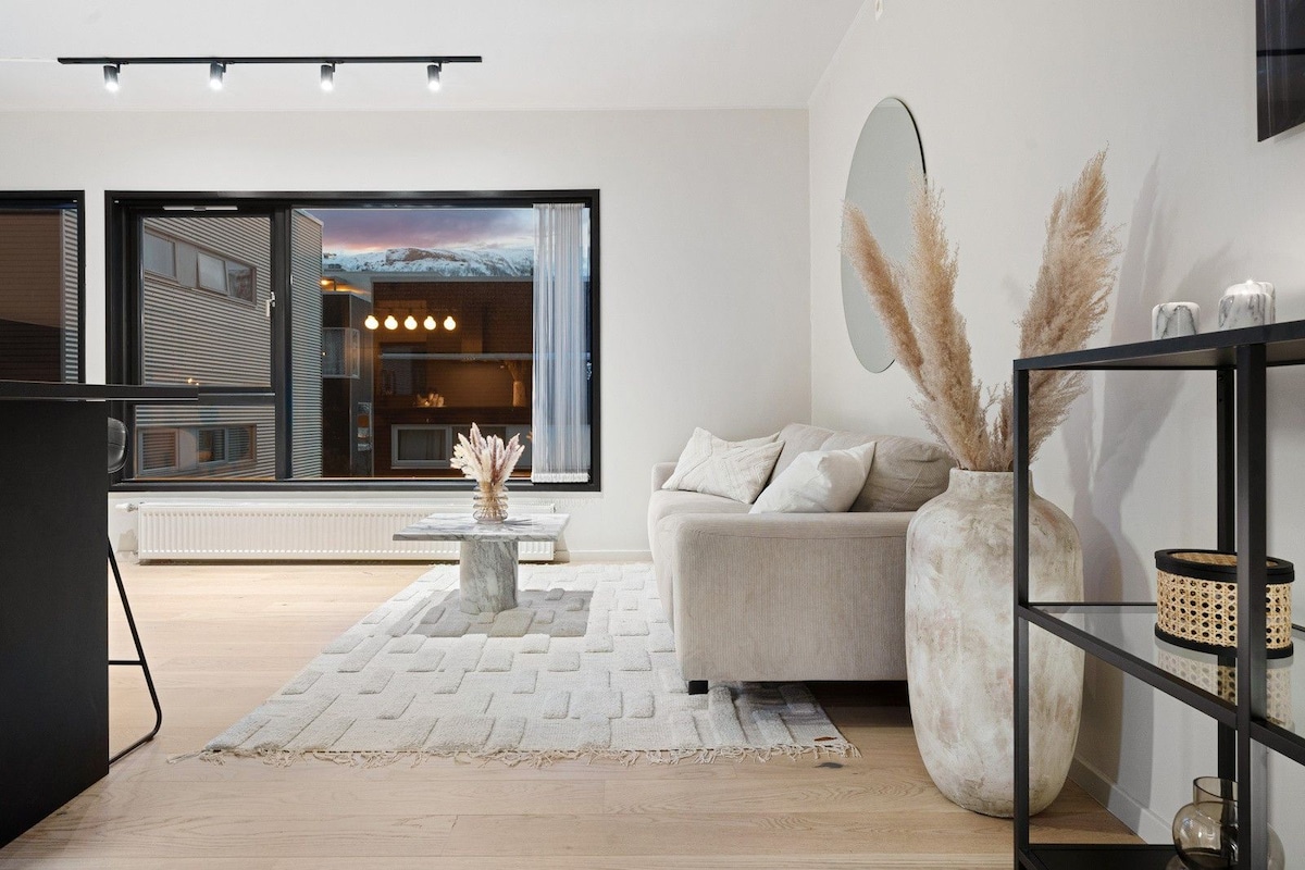 A light-filled living area is presented with a comfortable sofa and a minimalist coffee table. A decorative vase with pampas grass adds a natural touch. Large windows allow views of the outdoors, while aligned overhead lighting highlights the space.
