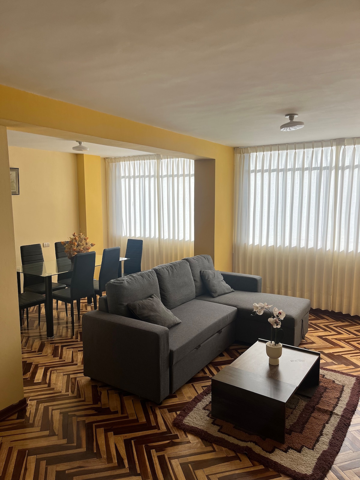 The living area features a comfortable gray sofa with decorative pillows, accompanied by a dark coffee table. Natural light filters through tall windows, highlighting the patterned hardwood floor. A dining area with four chairs is visible in the background, providing space for meals or gatherings.