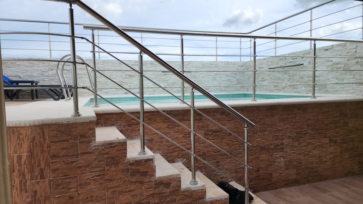 A well-maintained outdoor area features a small pool surrounded by a stone wall. A set of stairs with stainless steel railings leads up to the pool, offering safety and accessibility. Lounge chairs are positioned nearby, ready for relaxation.