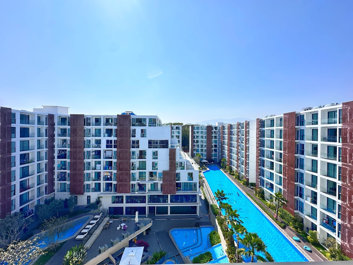 An aerial view of a contemporary apartment complex is displayed, featuring symmetrical white and red brick buildings surrounding a large turquoise pool. Lush greenery is visible along the pool's edge and in the courtyard area, enhancing the overall appeal of the community.