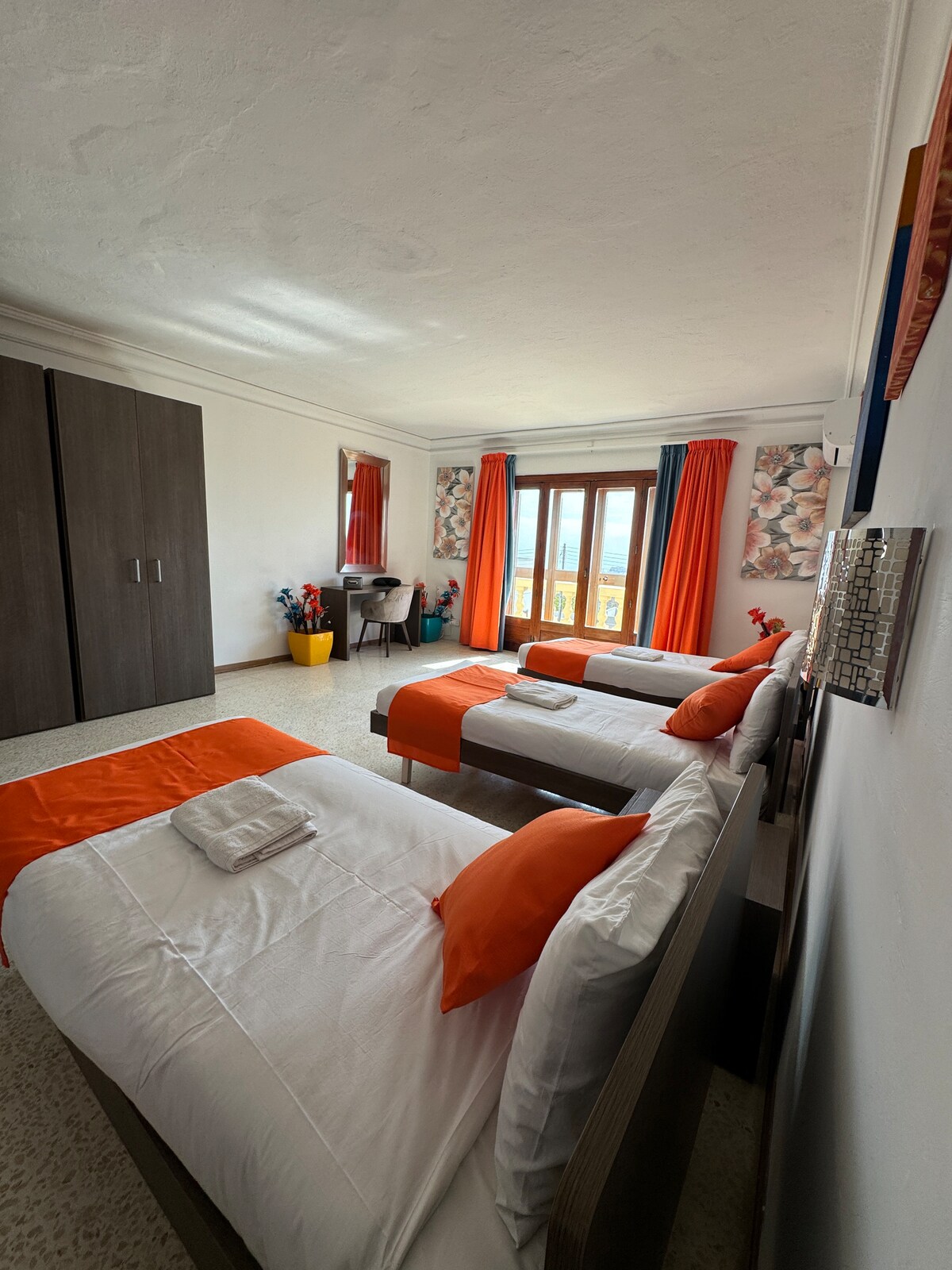 A spacious room features three neatly arranged beds adorned with bright orange accents. A large wardrobe stands against one wall, complemented by a wooden table and chairs near a window that allows natural light to fill the space. Floral wallpaper adds a subtle touch of color.