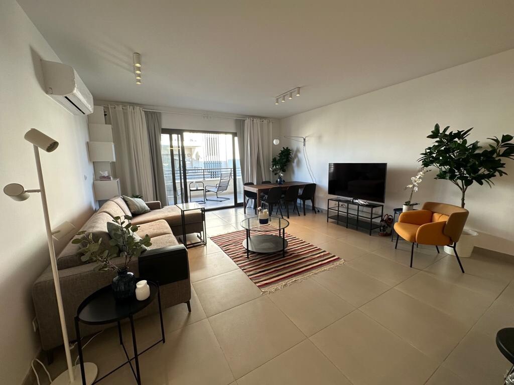 A bright living area is shown, featuring a comfortable sofa, a small side table, and a vibrant orange chair. A dining table accommodates six, while a television is positioned nearby. Large glass doors open to a balcony, enhancing the space with natural light.