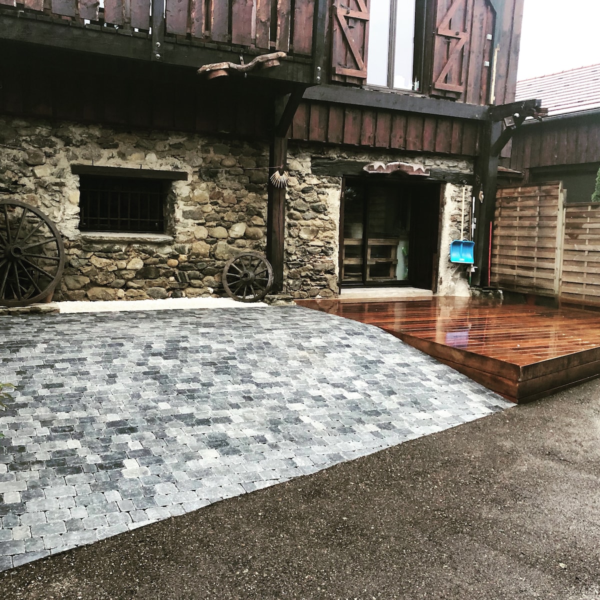 The exterior features a combination of stone and wood materials. A stone-paved path leads to a wooden deck area. Vintage wheels accentuate the scene, providing a rustic charm. Natural light highlights the textures of the building, with large windows visible in the background.