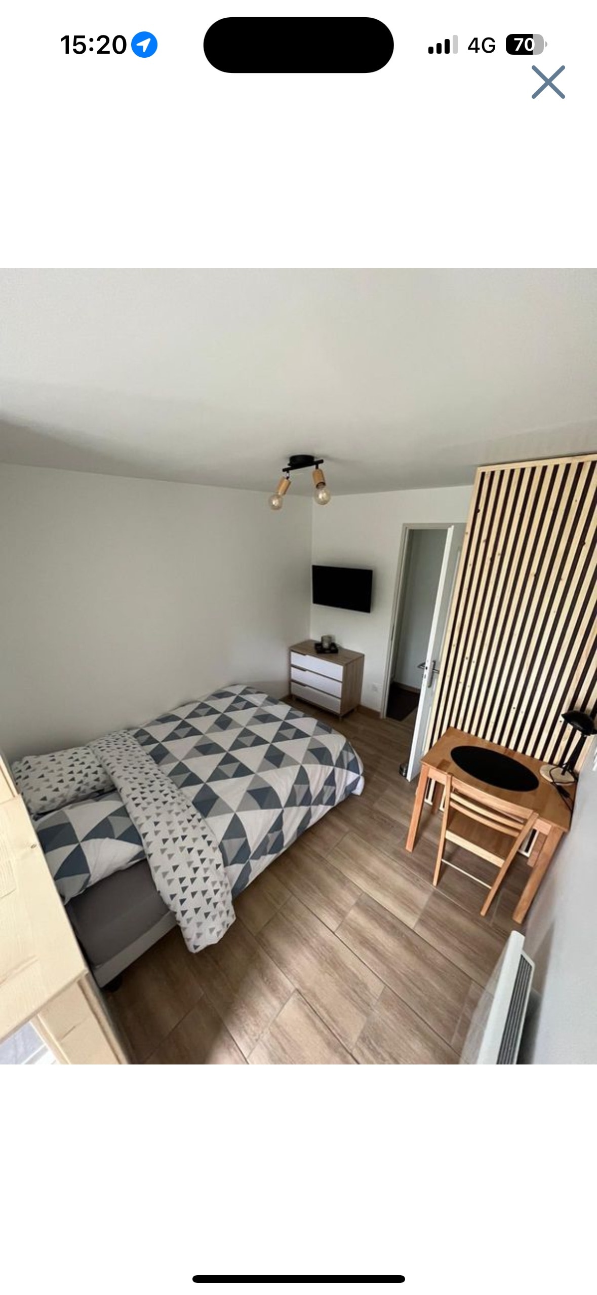 A cozy sleeping area is highlighted by a patterned bedding set on a spacious bed. A small wooden desk with a chair is present, along with a dresser and a wall-mounted TV. Natural light enters through a nearby door, enhancing the calm atmosphere.
