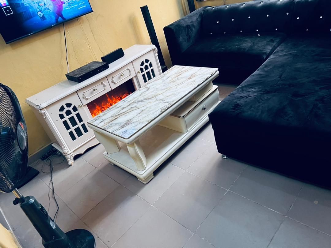 A black sectional sofa is positioned against the wall with decorative buttons, accompanied by a white coffee table featuring a marble top. An electric fireplace is visible beneath the television, and a standing fan is placed nearby, enhancing the comfort of the living area.