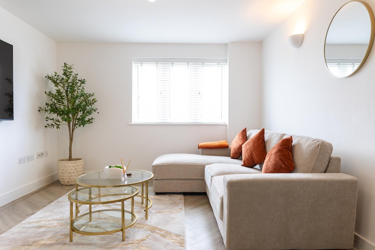 A modern living area features a beige sectional sofa with orange accent pillows, positioned beside a glass coffee table. A decorative plant is placed in a light-colored pot, while natural light filters through the window adorned with white shutters.