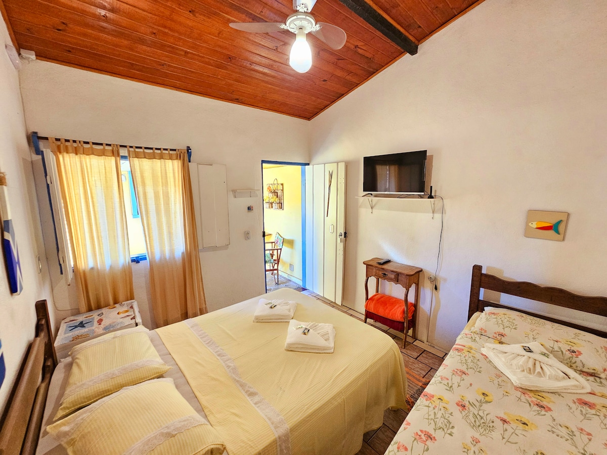 A bright sleeping area is presented with a spacious bed and a smaller bed indicated by decorative bedding. Natural light filters through sheer curtains, illuminating the wooden ceiling and providing a cozy ambiance. A small table and chair are visible, along with a wall-mounted television.