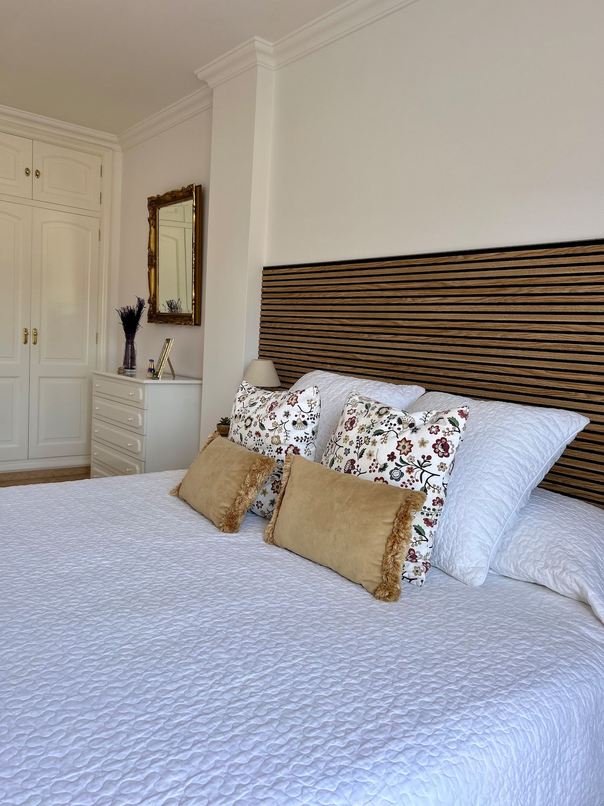 A spacious bedroom features a large bed with a textured white bedspread and decorative pillows. A wooden headboard adds warmth, while a tall wardrobe and dresser are visible, providing ample storage. Natural light brightens the room through a nearby window.