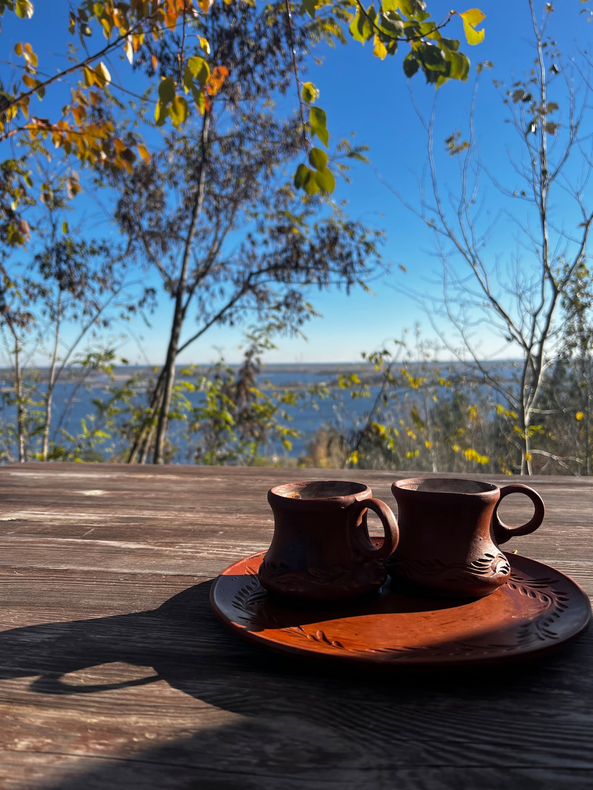 Two clay cups are placed on a rustic wooden table, accompanied by a round clay plate. In the background, a serene view of water and trees is visible under a clear blue sky. Soft shadows enhance the peaceful setting.