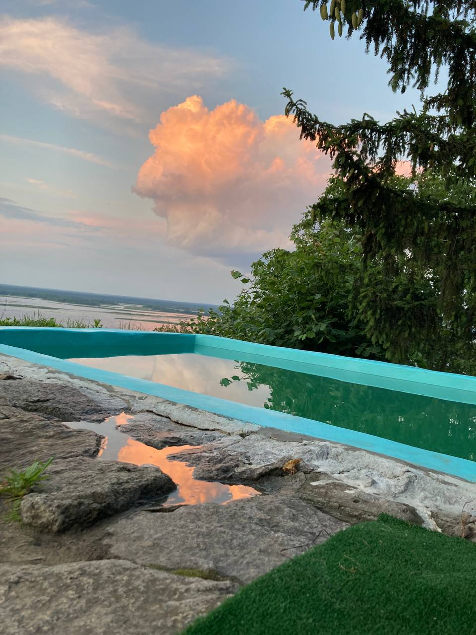 A calm pool reflects the sky, framed by natural stone that extends to the lush greenery nearby. The horizon is adorned with soft clouds tinged in orange, providing a serene backdrop to the tranquil water.