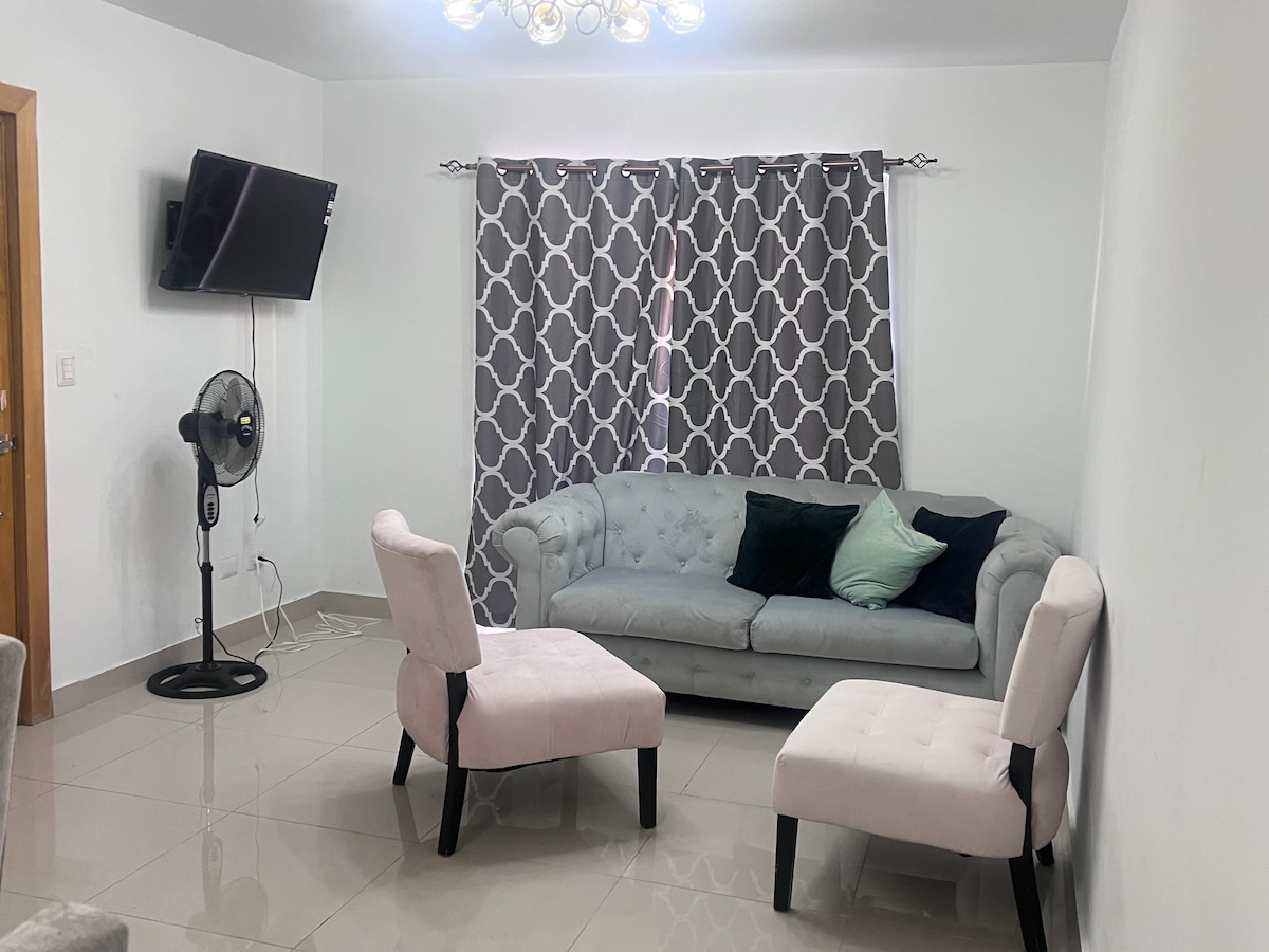 The living area features a light gray couch adorned with decorative pillows, accompanied by two soft pink chairs. A flat-screen television is mounted on the wall, and window treatments consist of patterned curtains that provide privacy while allowing soft light to filter through.