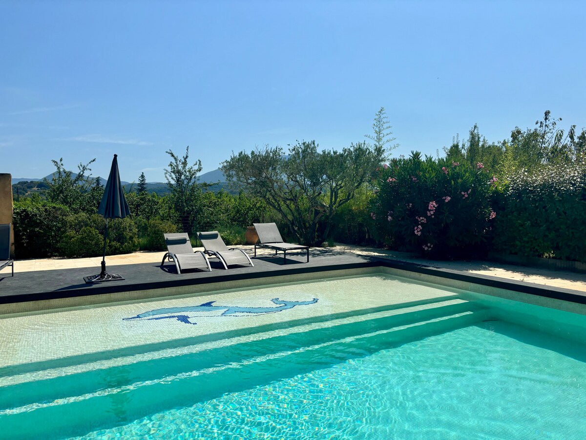 A serene swimming pool is featured, surrounded by lush greenery and blossoming plants. Comfortable lounge chairs are positioned beside the pool, with a shaded umbrella nearby. The clear blue sky is visible, enhancing the peaceful outdoor setting.