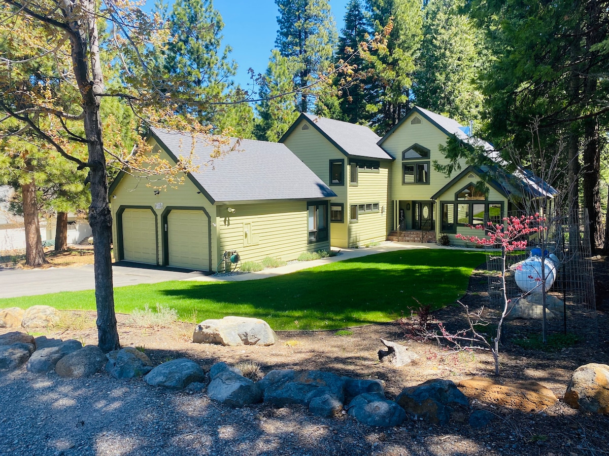 The exterior of a spacious green house is surrounded by tall trees and a manicured lawn. A gravel driveway leads to a two-car garage. The front features several large windows, allowing natural light to fill the interior. A tranquil atmosphere is created by the lush landscaping.