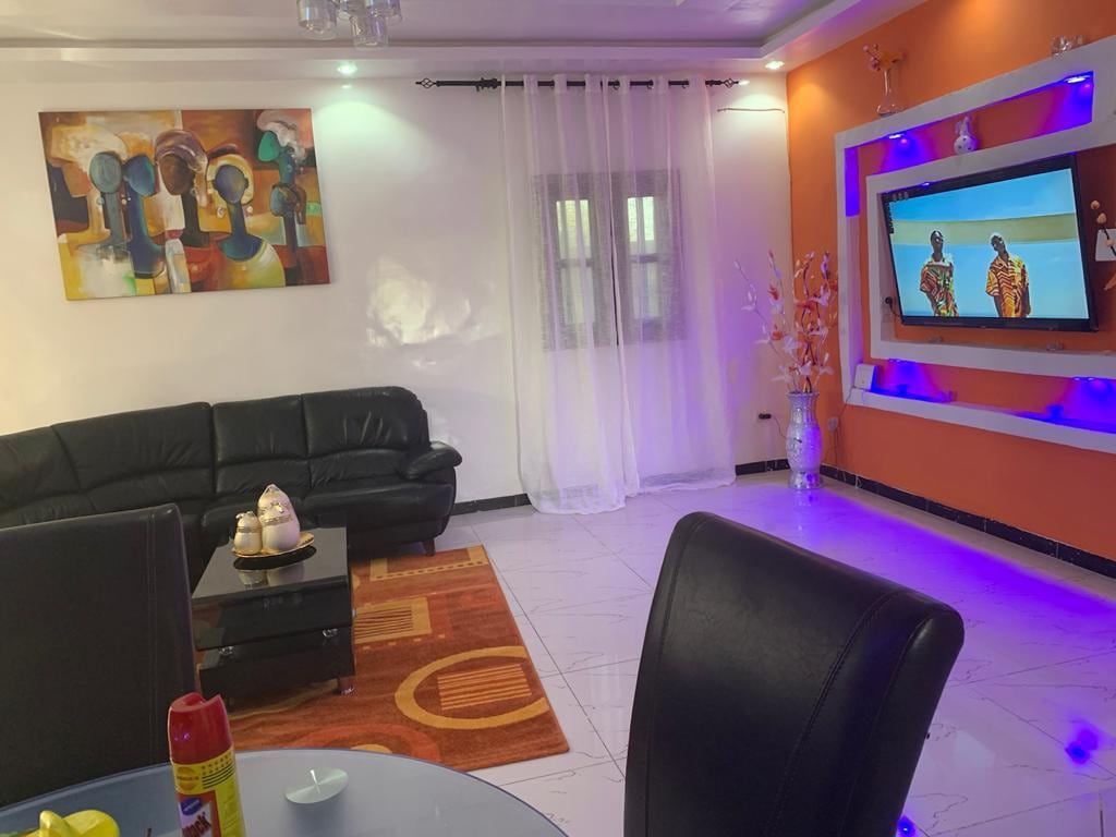 A modern living area is highlighted by a black leather sofa and a coffee table. Colorful artwork adorns the wall, while a flat-screen TV is mounted on a stylish display. Sheer curtains soften the natural light coming through a window, creating a vibrant yet relaxing space.
