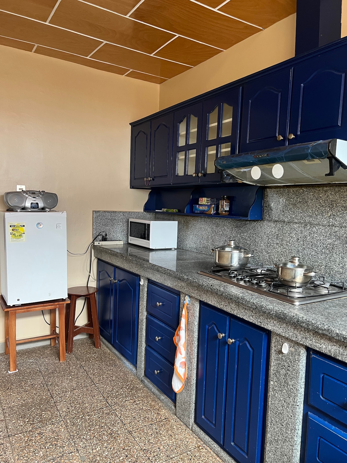 A functional kitchen is equipped with blue cabinetry and a countertop made of grey stone. Stainless steel pots are placed on a gas stove, while a microwave and a small refrigerator are visible. A wooden chair and an orange kitchen towel add a practical touch.
