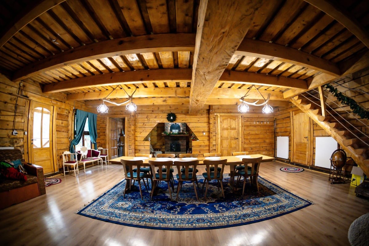 A spacious dining area features a long wooden table surrounded by chairs, set on a patterned area rug. The room is illuminated by natural light from large windows, enhancing the rustic wooden walls and ceiling beams. A cozy seating corner is visible, with a fireplace adding warmth.