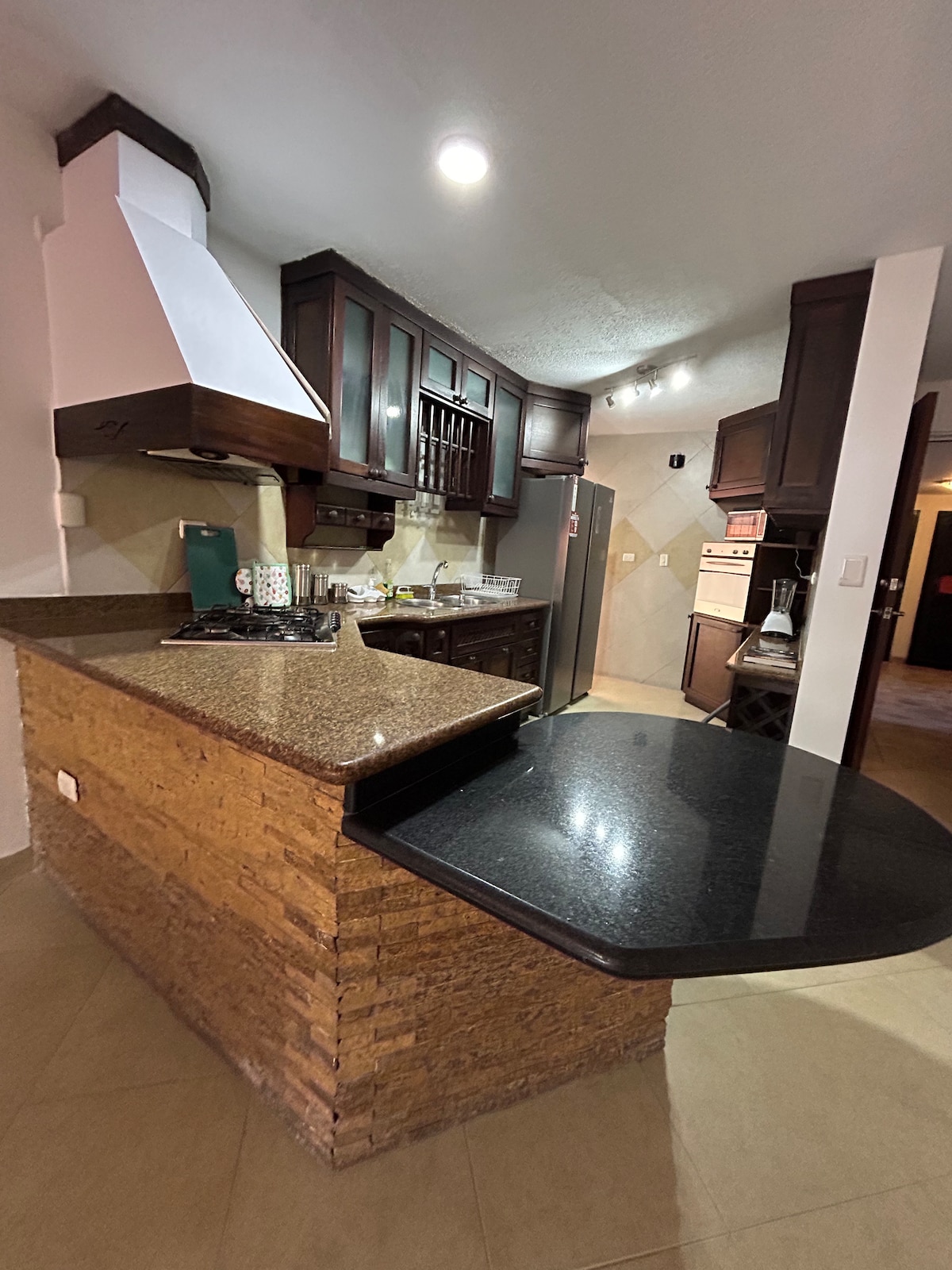The kitchen features dark wooden cabinets and a spacious granite countertop. A modern stovetop is integrated into the island, which also includes a breakfast bar. Natural light illuminates the area, highlighting the contemporary appliances and functional design.