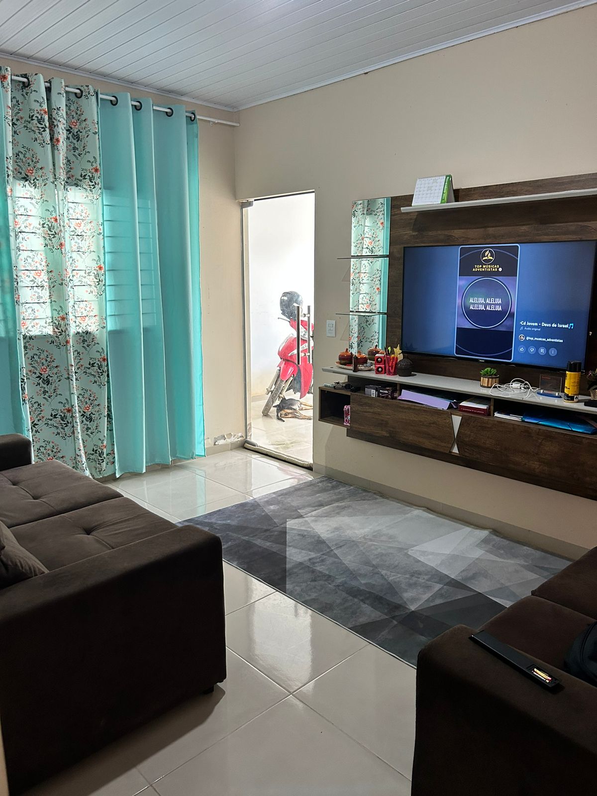 A comfortable living area features a dark sofa and a light-colored rug on tiled flooring. Floor-to-ceiling windows with sheer turquoise curtains allow natural light, while a television is mounted on a wooden entertainment unit. A door leads to an adjoining space.