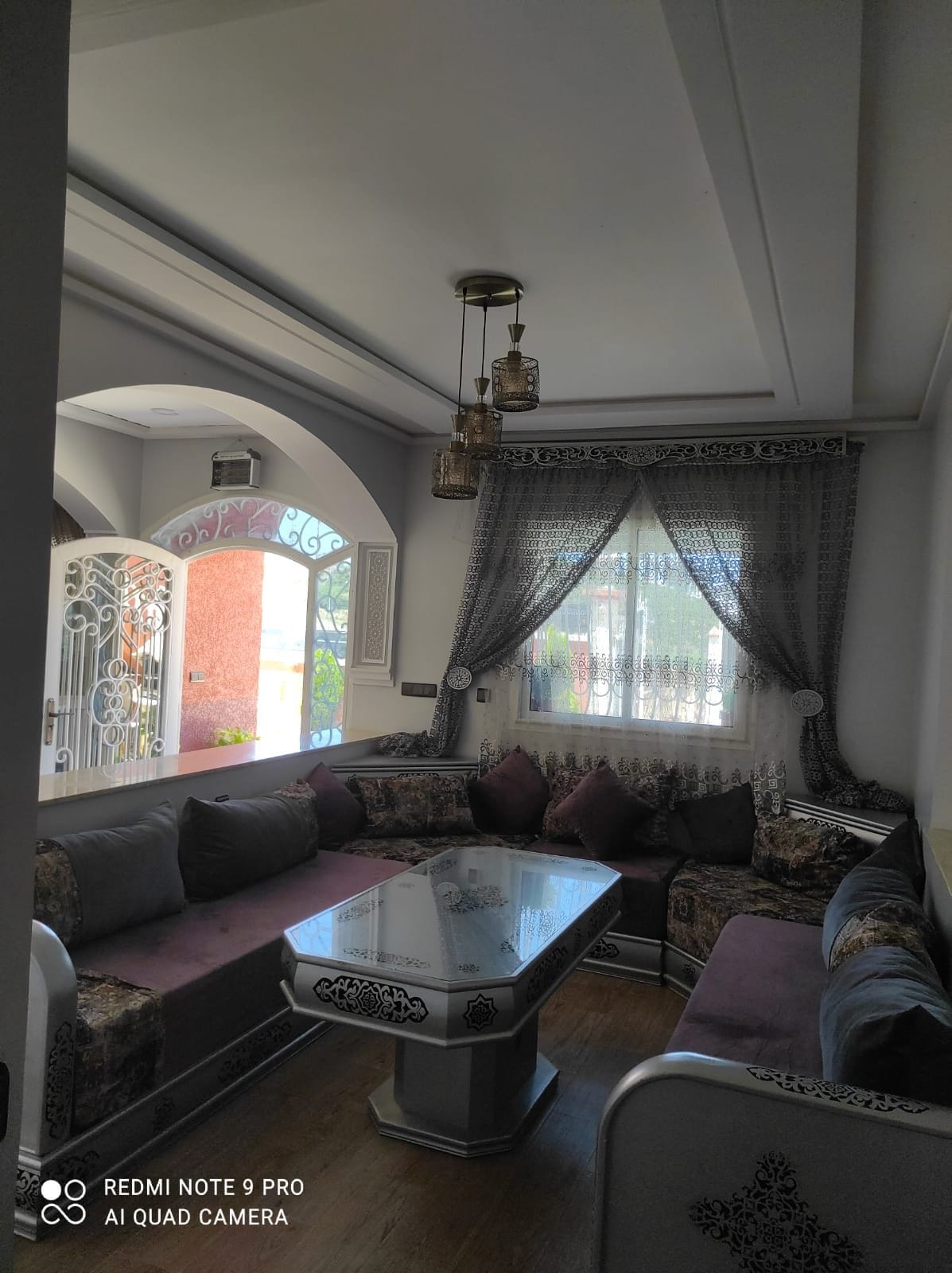 A comfortable living area features an ornate seating arrangement with plush cushions in shades of grey and purple. A glass table sits at the center, reflecting the natural light coming through the large window adorned with delicate curtains.
