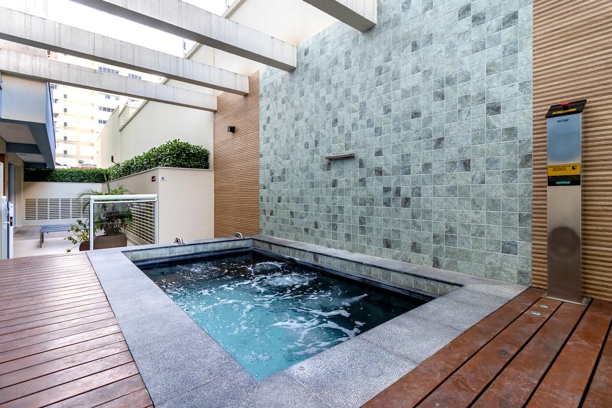 A tranquil spa area features a small jacuzzi surrounded by textured stone walls and wooden decking. Water bubbles gently in the jacuzzi, providing a peaceful atmosphere. Greenery is visible above, adding a touch of nature to the space.
